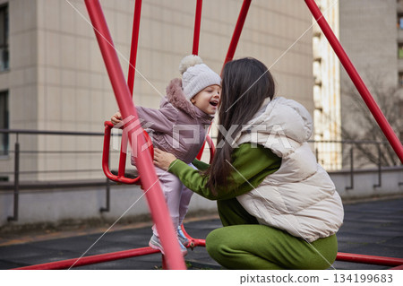 Mother pushing happy daughter on a swing set in an urban playground 134199683