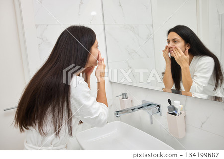 woman applying skincare cream to her face in the bathroom mirror woman applying skincare cream to her face in the bathroom mirror 134199687