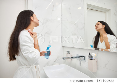 Woman cleansing her face with toner and cotton pad looking in bathroom mirror Woman cleansing her face with toner and cotton pad looking in bathroom mirror 134199688