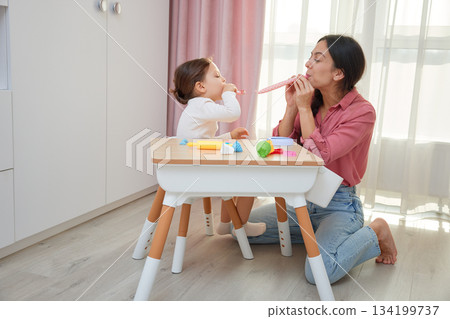 Mother and daughter celebrating and playing with party blowers at a small table 134199737
