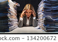 A businesswoman with a sad expression on her face, surrounded by a huge pile of documents, holding her head 134200672