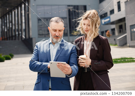 Confident businesspersons standing in front of modern office building. Man and woman talking and looking at documents. Banking and financial market concept. 134200709