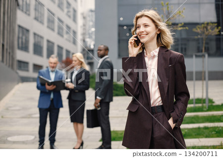 Close up on caucasian woman wearing brown jacket and talking on the phone. Confident businesspeople standing in front of modern office building. Men and women talking and looking at documents. 134200711
