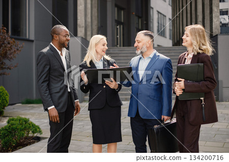 Confident businesspersons standing in front of modern office building. Men and women talking and holding documents. Banking and financial market concept. Confident businesspersons standing in front of modern office building. Men and women talking and holding documents. Banking and financial market concept. 134200716