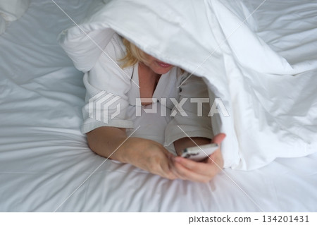 Smiling woman lying on a bed with a blanket over her head while using the mobile phone 134201431