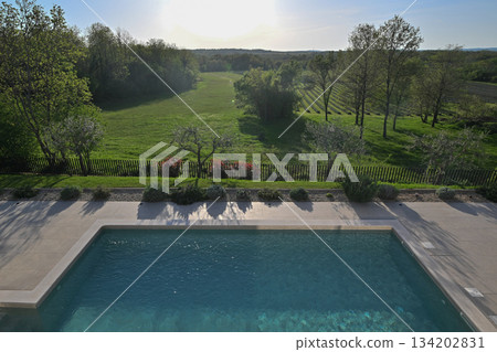 Elevated view of a modern swimming pool with clear blue water, overlooking a picturesque, expansive green landscape of fields, trees, and vineyards under a bright sky. Perfect for tranquil luxury 134202831