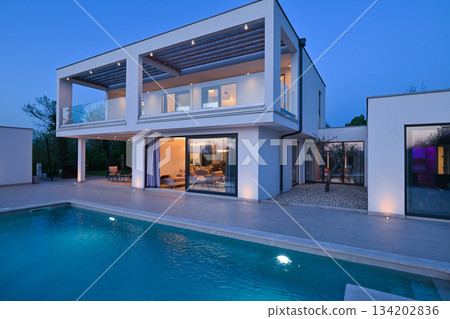 Exterior view of a modern luxury house with a swimming pool at twilight. Illuminated two-story white villa with large glass windows, patio, terrace, and glowing blue water 134202836