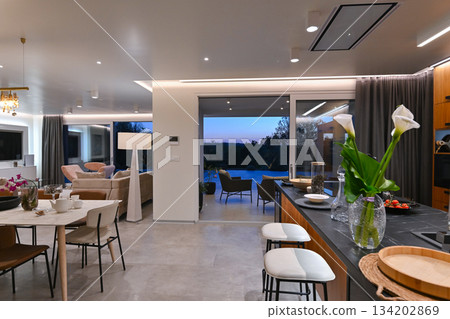 Modern luxury living room interior at sunset. Open-plan space featuring beige sofas, marble dining table, large glass windows with sun flare, and view of a patio. Stylish contemporary home design 134202869