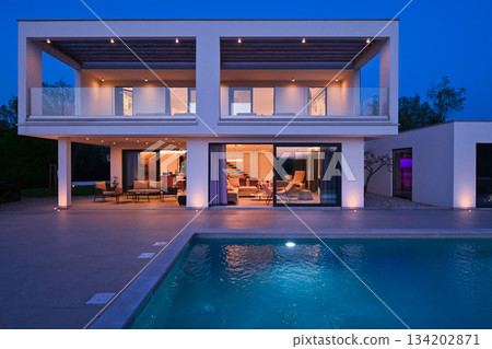 Exterior view of a modern luxury house with a swimming pool at twilight. Illuminated two-story white villa with large glass windows, patio, terrace, and glowing blue water 134202871
