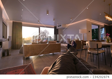 Modern luxury living room interior at sunset. Open-plan space featuring beige sofas, marble dining table, large glass windows with sun flare, and view of a patio. Stylish contemporary home design Modern luxury living room interior at sunset. Open-plan space featuring beige sofas, marble dining table, large glass windows with sun flare, and view of a patio. Stylish contemporary home design 134202874