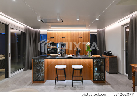 Modern luxury kitchen interior with wooden cabinetry, central island with bar stools, and ambient LED lighting. Stylish contemporary design with large windows and dark countertops at twilight Modern luxury kitchen interior with wooden cabinetry, central island with bar stools, and ambient LED lighting. Stylish contemporary design with large windows and dark countertops at twilight 134202877