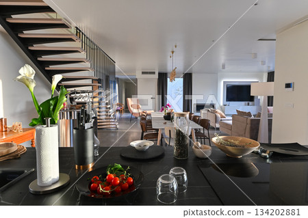 Modern luxury open-plan interior featuring a floating staircase, marble dining table, wooden kitchen, and beige sofa. Spacious contemporary home design with elegant lighting and gray tiled floors Modern luxury open-plan interior featuring a floating staircase, marble dining table, wooden kitchen, and beige sofa. Spacious contemporary home design with elegant lighting and gray tiled floors 134202881