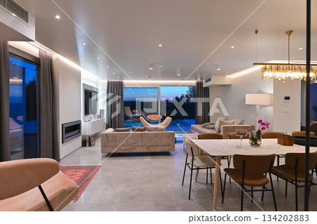 Modern luxury living room interior at sunset. Open-plan space featuring beige sofas, marble dining table, large glass windows with sun flare, and view of a patio. Stylish contemporary home design 134202883