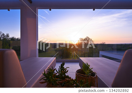 Sunset view from a luxurious modern patio with lounge chairs, overlooking a tranquil green landscape of trees and vineyards, perfect for a relaxing evening 134202892