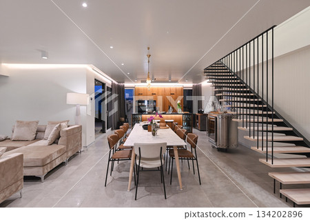 Modern luxury open-plan interior featuring a floating staircase, marble dining table, wooden kitchen, and beige sofa. Spacious contemporary home design with elegant lighting and gray tiled floors 134202896