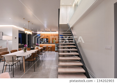 Modern luxury open-plan interior featuring a floating staircase, marble dining table, wooden kitchen, and beige sofa. Spacious contemporary home design with elegant lighting and gray tiled floors 134202903