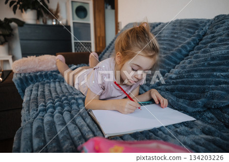 Girl drawing in a notebook while relaxing on a blue couch in a cozy living room Girl drawing in a notebook while relaxing on a blue couch in a cozy living room 134203226