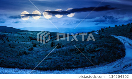 country dirt road through mountains on a summer night. scenic view of rolling hills under dark sky with clouds in full moon light. beautiful alpine meadow for finance investment or abstract concepts 134204502