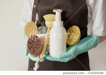 Person wearing gloves and apron holding natural sponges, bamboo brush, and soap bottles for cleaning 134204590