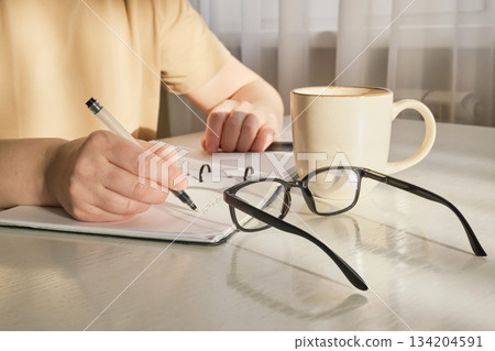 Person writing ideas, planning, or studying at desk with coffee and reading glasses nearby 134204591