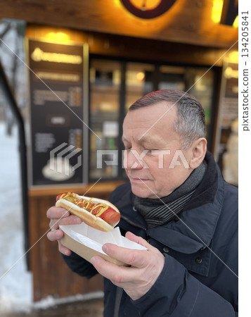 Adult man holding hot dog near street food kiosk in winter. Outdoor city scene with warm light. Adult man holding hot dog near street food kiosk in winter. Outdoor city scene with warm light. 134205841