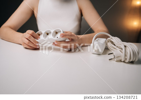 Cropped shot of unrecognizable woman gripping white triple electrical socket sitting at white desk, highlighting electrical work, home improvement, energy efficiency in dark interior setting 134208721