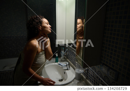 Middle aged curly haired woman, standing naked wrapped in bath towel in front of a bathroom mirror, touching her face while admiring her reflection during daily morning beauty and hygiene procedures Middle aged curly haired woman, standing naked wrapped in bath towel in front of a bathroom mirror, touching her face while admiring her reflection during daily morning beauty and hygiene procedures 134208729