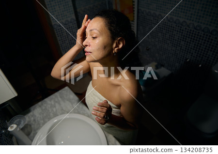 Confident portrait of a middle aged pretty woman applying cosmetic product, standing in front of a bathroom mirror, wrapped in bath towel, enjoying daily morning routine and procedures 134208735