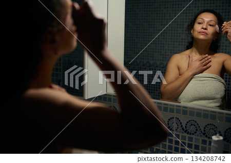 Reflection in the mirror of a young adult woman performing face lifting treatment using a roller stone massager during everyday beauty spa procedures, standing in the bathroom wrapped in a bath towel Reflection in the mirror of a young adult woman performing face lifting treatment using a roller stone massager during everyday beauty spa procedures, standing in the bathroom wrapped in a bath towel 134208742