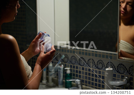 Close-up woman holing natural crystal antiperspirant from alum salts, standing at bathroom sink, and looking at her reflection in the mirror. Eco-friendly and organic cosmetics for health and wellness Close-up woman holing natural crystal antiperspirant from alum salts, standing at bathroom sink, and looking at her reflection in the mirror. Eco-friendly and organic cosmetics for health and wellness 134208751