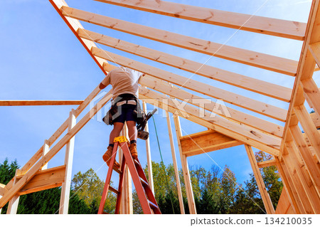 Worker builds wooden roof frame on while standing on ladder in construction site Worker builds wooden roof frame on while standing on ladder in construction site 134210035