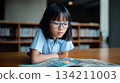 Young girl with glasses is deeply engaged in reading a colorful comic book at a wooden table in a library, surrounded by shelves filled with books and educational materials 134211003