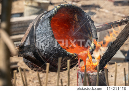 Blacksmith holding a hot crucible by the iron pliers from furnace and pouring the melting gold into the statue block. The process to making of metal statue in local Thailand traditional. Blacksmith holding a hot crucible by the iron pliers from furnace and pouring the melting gold into the statue block. The process to making of metal statue in local Thailand traditional. 134211458