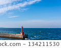 A red lighthouse (beacon) stands at the end of a breakwater stretching out into the sea under blue skies and clouds. 134211530