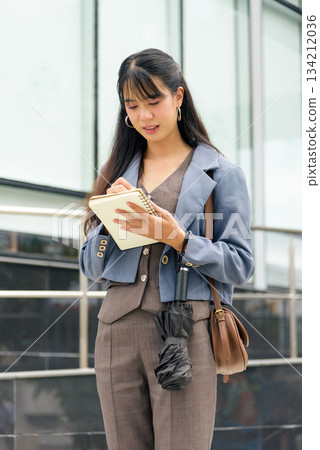 Young woman on city street, business professional lifestyle person working outdoor writing notes in book carrying umbrella shoulder bag standing urban city background focused on task. 134212036
