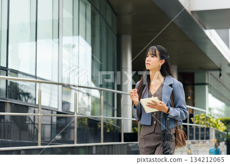 Asian woman walks street outdoor, engaged in business work lifestyle activity, considering travel plans, holding notebook and pen, showcasing professional look near impressive urban architecture. 134212065
