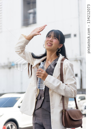 Travel woman person on city street business work outdoor lifestyle, shielding eyes from harsh sun, feeling hot summer weather, holding bottle water for hydration. 134212073