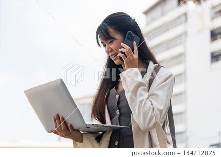 Smiling young Asian woman, business person, on busy city street handles work with laptop, communicating on phone, managing professional urban lifestyle outdoors, embodying modern travel spirit. 134212075