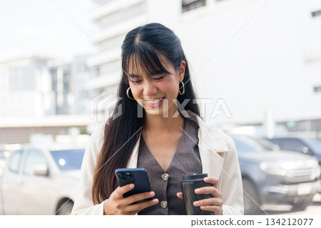 Smiling young business woman, engaged person enjoying modern urban lifestyle, using smartphone, holding coffee cup, walking outdoor city street, representing work and travel. 134212077