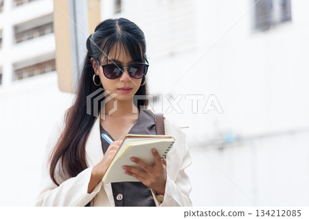 Business woman, person on city street enjoying outdoor work lifestyle, stylish young professional diligently takes notes, planning future travel goals. Business woman, person on city street enjoying outdoor work lifestyle, stylish young professional diligently takes notes, planning future travel goals. 134212085