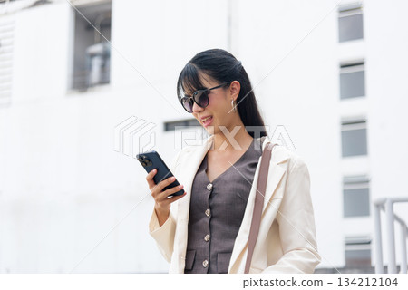 Professional business woman, stylish person, actively uses smartphone on urban street, enjoying outdoor work travel lifestyle, capturing contemporary people commute. 134212104