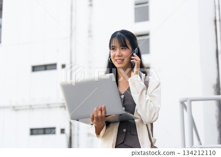 Smiling happy Asian woman for business work outdoor on street, talking on mobile phone, using laptop, representing modern urban city lifestyle for young professionals. Smiling happy Asian woman for business work outdoor on street, talking on mobile phone, using laptop, representing modern urban city lifestyle for young professionals. 134212110
