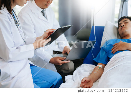 Two doctors talking to a patient lying in his bed with receiving saline solution Two doctors talking to a patient lying in his bed with receiving saline solution 134213352
