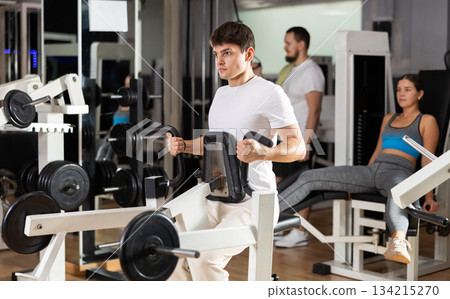 Guy in gym performs exercises for arm muscles using simulators and fitness center equipment. Guy in gym performs exercises for arm muscles using simulators and fitness center equipment. 134215270