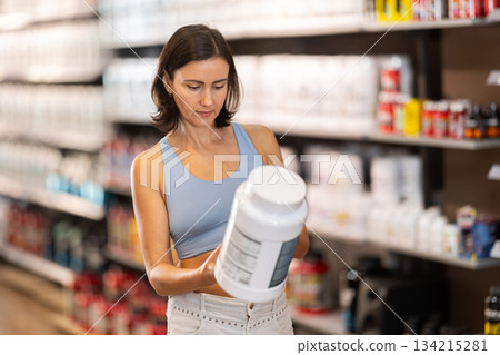 Focused woman choosing protein powder jar in sports nutrition store 134215281