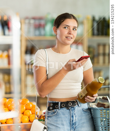 European young woman customer at shop scans QR code on peach juice bottle using phone camera 134215295