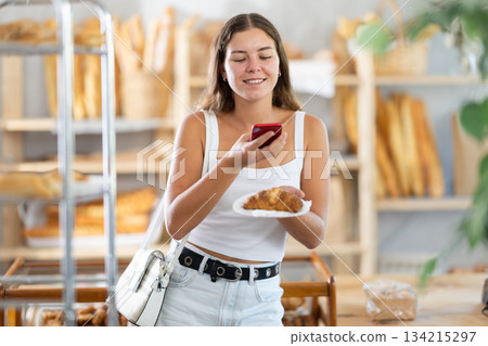 Young woman stands in bakery, scanning a croissant with her phone Young woman stands in bakery, scanning a croissant with her phone 134215297