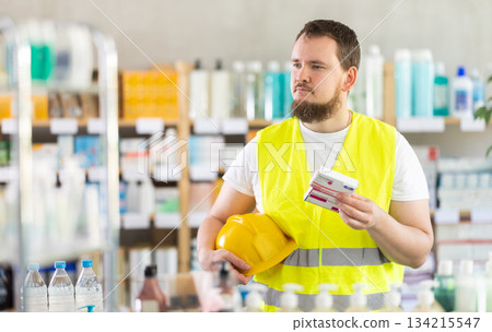 Male builder in yellow vest with helmet in his hands buys paracetamol medicine at pharmacy Male builder in yellow vest with helmet in his hands buys paracetamol medicine at pharmacy 134215547