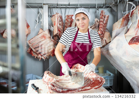 Positive female butcher cutting veal ribs in cold storage room of store 134216332