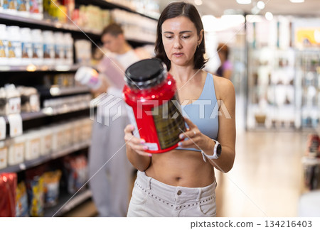 Focused woman choosing protein powder jar in sports nutrition store Focused woman choosing protein powder jar in sports nutrition store 134216403